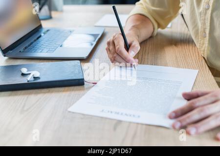Beschnittene Ansicht des Geschäftsmannes, der auf einem verschwommenen Vertrag neben einem Notebook und einem Kopfhörer auf dem Tisch schrieb, Stockbild Stockfoto