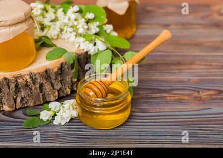 Süßes Honigglas umgab die Akazienblüten des Frühlings. Honig fließt aus einem Löffel in ein Glas. Gläser von klarem frischen Akazienhonig auf Holzgrund. Stockfoto