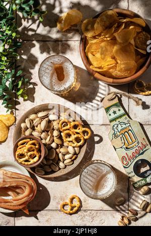 Bier-Party-Konzept im Freien mit Bier in Gläsern, Snacks, Chips, Nüssen, Brezeln auf Fliesenhintergrund, Draufsicht, Flat Lay Stockfoto