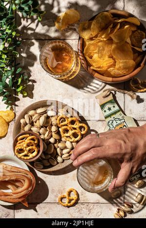 Bier im Freien Party Konzept. Die Hand des Mannes nimmt ein Glas Bier vom Tisch mit verschiedenen Snacks. Stillleben auf Fliesenhintergrund mit harten Schatten, zu Stockfoto