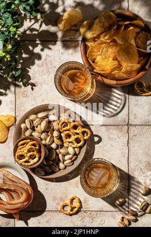 Sommerbier-Party im Freien. Zwei Gläser Bier mit verschiedenen Snacks, Chips, Nüssen, Brezeln auf Fliesenhintergrund mit harten Schatten, Draufsicht, fl Stockfoto
