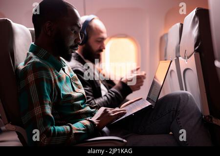 Geschäftsmann mit Sitz, der in der Economy-Klasse auf Geschäftsreise mit dem Flugzeug ist und während des Sonnenuntergangs auf einem Laptop arbeitet. Männlicher Mitarbeiter, der mit einem Handelsflug bei einer internationalen Fluggesellschaft fliegt. Stockfoto