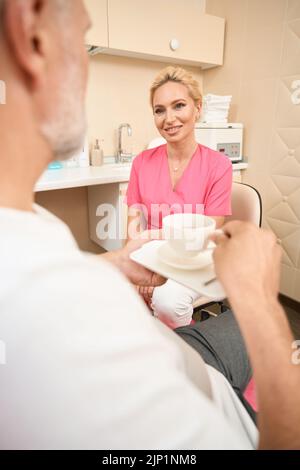 Lächelnde Kosmetologin, die den Patienten beim Kaffeetrinken im Kosmetologiebüro berät Stockfoto