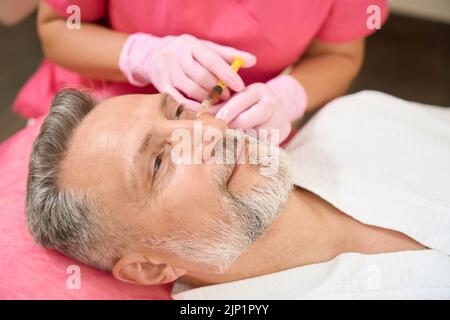 Hände einer Kosmetikerin, die dem grauhaarigen Mann unter dem Auge eine Schönheitsspritze zuführt Stockfoto