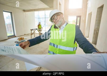 Der männliche Vorarbeiter hält eine erweiterte Zeichnung und spricht auf dem Mobiltelefon Stockfoto