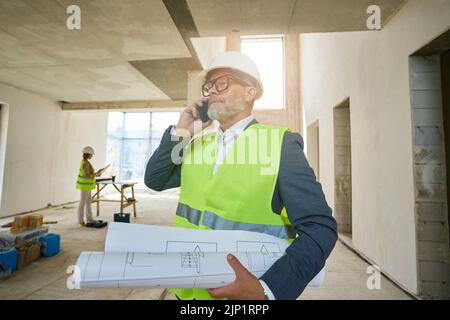 Vorarbeiter hält gefaltete Zeichnungen und spricht auf dem Mobiltelefon Stockfoto