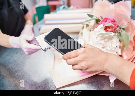 Frau hält Telefon in der Nähe des Terminals, um für den bestellten Blumenstrauß in der Blumenboutique zu bezahlen Stockfoto
