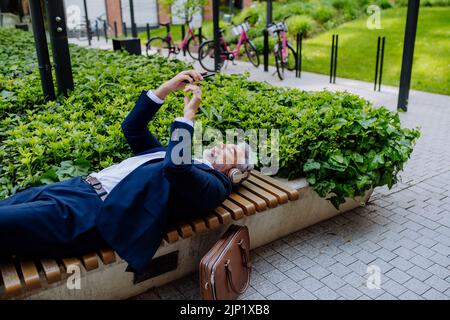 Ein reifer Geschäftsmann, der in Kopfhörern Musik genießt und sich während der Arbeitspause auf der Bank im Stadtpark entspannt. Stockfoto