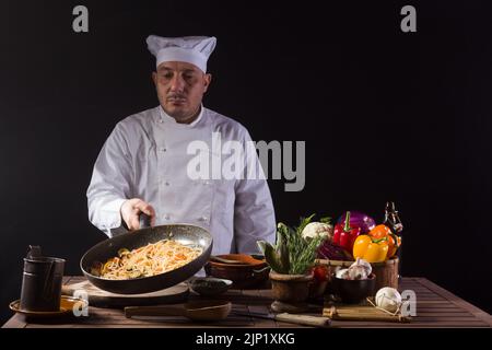 Chefkoch in weißer Uniform hält eine Bratpfanne und sautiert Spaghetti mit frischem Gemüse, das in der Luft fliegt, bevor er während der Arbeit in einem Restaurant serviert Stockfoto