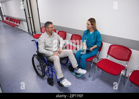 Optimistischer Patient im Rollstuhl kommuniziert mit einem freundlichen behandelnden Arzt in der Lobby einer Ambulanz Stockfoto