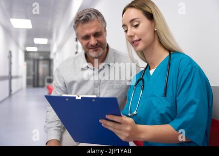 Europäische Ärztin mit langen Haaren, die Dokumenten für einen schönen männlichen Patienten zeigt Stockfoto