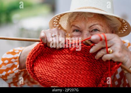 Porträt einer älteren Frau, die draußen sitzt und einen roten Schal strickt. Stockfoto