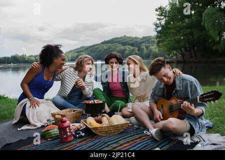 Eine Gruppe von Freunden, die Spaß beim Picknick in der Nähe eines Sees haben, auf der Decke sitzen und Gitarre spielen. Stockfoto