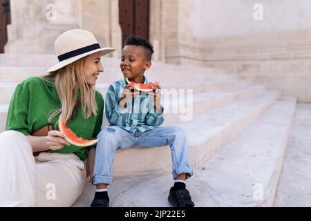 Multirassischer Sohn mit Mutter, der an heißen sonnigen Tagen Wassermelone auf der Straße isst, Reisekonzept für Sommerferien. Stockfoto