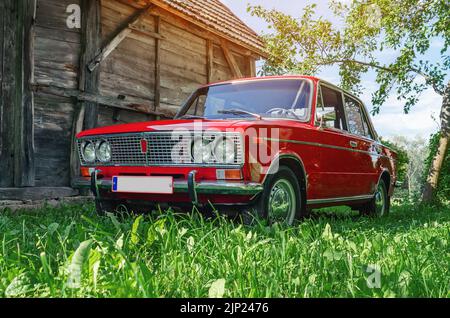Nahaufnahme des roten Oldtimers Lada auf dem Land. Alte hölzerne Scheune Hintergrund. Selektiver Fokus von grünem Gras, das durch Sonnenlicht beleuchtet wird. Stockfoto