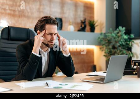 Kopfschmerzen, Migräne. Ermüdungskaukasischer Geschäftsmann, Büroangestellter, sitzt an einem Arbeitsplatz in einem modernen Büro, berührt seine Schläfen, massiert den Kopf, fühlt sich gestresst, überarbeitet, Brauchen Ruhe und Kaffee-Brak Stockfoto