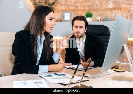 Kooperationskonzept. Zwei erfolgreiche kaukasische Mitarbeiter, Geschäftspartner, sitzen an einem Tisch im Büro, schauen auf den Computerbildschirm, studieren konzentriert die finanziellen Aussichten und Risiken des Unternehmens Stockfoto