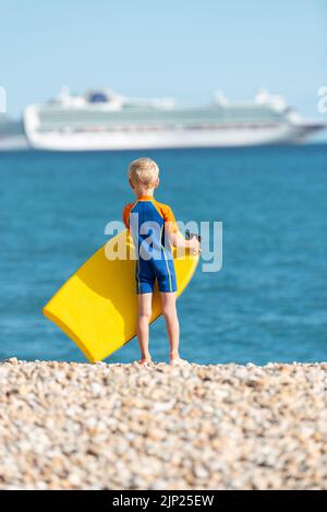 Der junge blonde Junge in einem blauen Neoprenanzug steht an einem Kiesstrand und hält ein gelbes Bodyboard mit Blick auf das Meer und ein Kreuzschiff in der Ferne Stockfoto