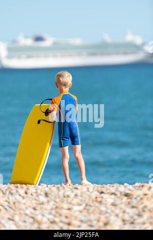 Der junge blonde Junge in einem blauen Neoprenanzug steht an einem Kiesstrand und hält ein gelbes Bodyboard mit Blick auf das Meer und ein Kreuzschiff in der Ferne Stockfoto