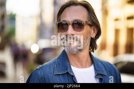 Glücklicher älterer Mann auf der Straße der europäischen Stadt in Sonnenbrille und Denim-Hemd. Grau bärtig mittleren Alters schöner Freiberufler Mann lächelnd Blick seitwärts tragen lässig. Reisekonzept. Stockfoto