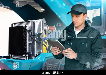 Servicearbeiter mit digitalem Tablet auf dem Hintergrund des Traktors mit offener Motorhaube Stockfoto