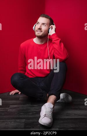 Eine vertikale Aufnahme eines Mannes, der in Kopfhörern auf dem Boden sitzt und Musik hört und einen roten Pullover trägt Stockfoto