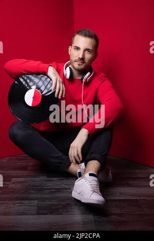 Ein Mann in einem roten Pullover sitzt auf dem Boden mit weißen Kopfhörern, die seinen Arm auf einem schwarzen Vinyl aufsetzen Stockfoto