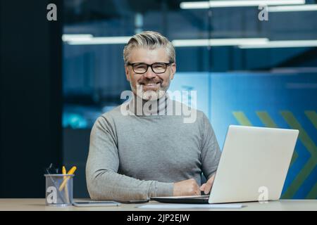 Porträt eines erfolgreichen Geschäftsmannes in einem modernen Büro, eines älteren, grauhaarigen Mannes mit Brille, lächelnd und mit Blick auf die Kamera, eines Investors, der am Laptop arbeitet Stockfoto