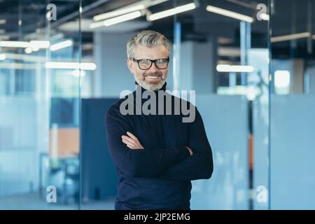 Porträt eines erfolgreichen Geschäftsmannes im modernen Büro mit gekreuzten Armen, grauhaarigen männlichen Investor lächelnd und Blick auf die Kamera, Chef in Brille und Jeans Stockfoto