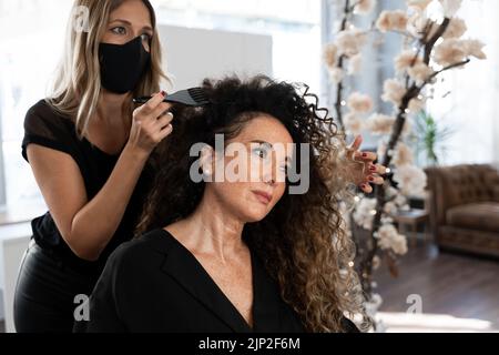 Friseur mit Maske Kämmen und das lockige Haar eines Kunden in einem Salon zu arrangieren Stockfoto