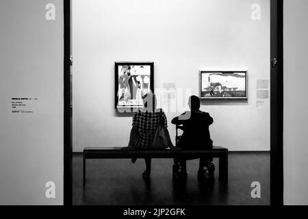 Eine Graustufenaufnahme eines alten Paares, das zusammen sitzt und abstrakte Gemälde in einem Museum bewundert Stockfoto