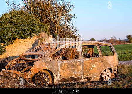 Ein altes verlassenes Auto, das auf einem Feld in der Nähe einer Asphaltstraße ruiniert wurde Stockfoto