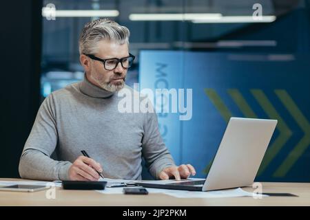 Seriöser Geschäftsmann hinter der Papierarbeit im Büro, grauhaarige Anleger, die Dokumente unterzeichnen, Mann, der im modernen Büro mit Konten und Laptop arbeitet, grauhaariger und reifer Geldgeber Stockfoto