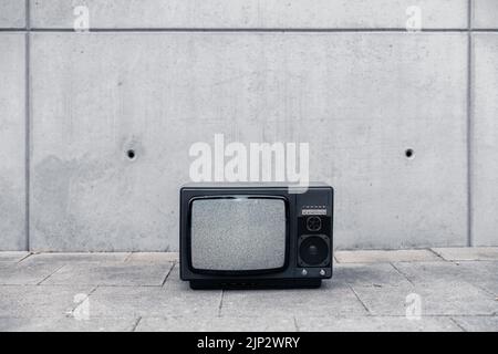 Ein alter Retro-fernseher stand draußen auf der Straße vor einer schmuddeligen Betonwand ohne Signal auf dem Bildschirm, nur Lärm Stockfoto