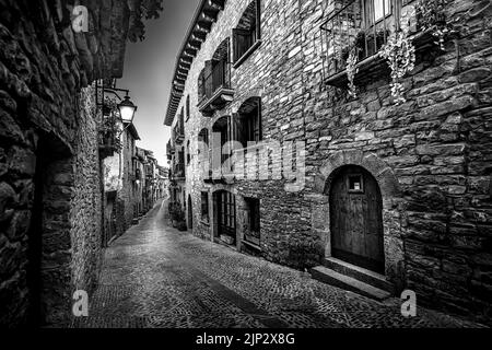 Schwarz-weiße mittelalterliche Altstadt mit Steinhäusern und Kopfsteinpflasterboden, Straßenlampen und dunkler und nächtlicher Atmosphäre. Ainsa, Spanien. Stockfoto