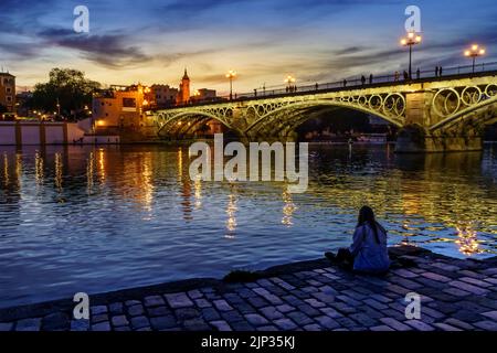 Triana-Brücke in Sevilla über den Guadalquivir-Fluss bei Sonnenuntergang, mit Lichtern, die sich im Flusswasser spiegeln, eine idyllische und charmante Szene. Andalusien. Stockfoto