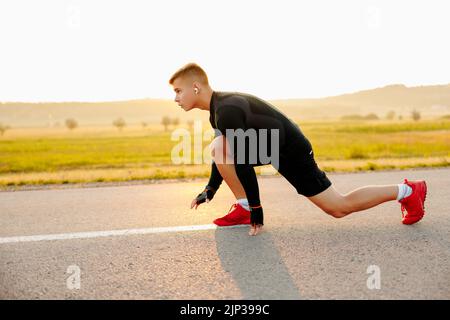 Nahaufnahme des männlichen Crouch Start auf Asphaltspur draußen und Drehen zurück zur Kamera. Er beugt sich am Körper und legt die Hände auf die Straße, während er fühlt Stockfoto
