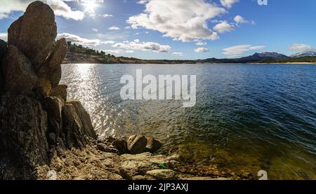 Landschaft aus See und Felsen mit der Sonne durch die Wolken, blauer Himmel und Reflexionen im Wasser. Stockfoto