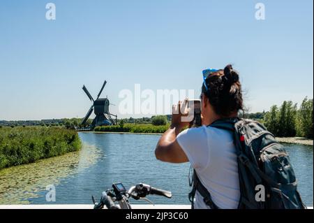 Kinderdijk, Niederlande. 13. August 2022. Man sieht einen Mann, der mit seinem Mobiltelefon ein Foto von einer der Windmühlen macht. In der schönen, wasserreichen Gegend in der Nähe von Dordrecht finden Sie die Windmühlen von Kinderdijk. Diese 19 prächtigen Windmühlen, die um 1740 erbaut wurden, stehen dort als Teil eines größeren Wassermanagementsystems, um Überschwemmungen zu verhindern. Heute symbolisieren sie die niederländische Wasserwirtschaft und wurden 1997 zum UNESCO-Weltkulturerbe erklärt. Jedes Jahr besuchen Tausende von Touristen aus der ganzen Welt diesen ikonischen Ort. Kredit: SOPA Images Limited/Alamy Live Nachrichten Stockfoto