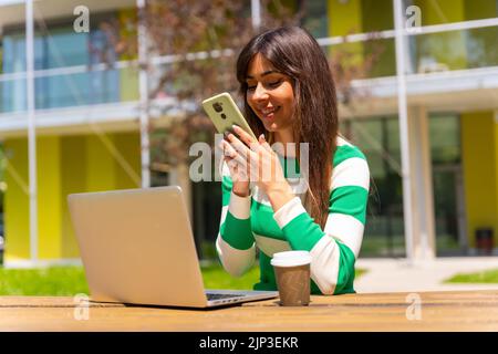 Eine kaukasische Frau in grün-weiß gestreifter Bluse, die mit einem Computer vor einem Tisch in der Natur arbeitet und die Fotos am Telefon überprüft Stockfoto