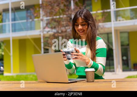 Eine kaukasische Frau in grün-weiß gestreifter Bluse, die mit einem Computer vor einem Tisch in der Natur arbeitet und die Fotos vor der Kamera überprüft Stockfoto