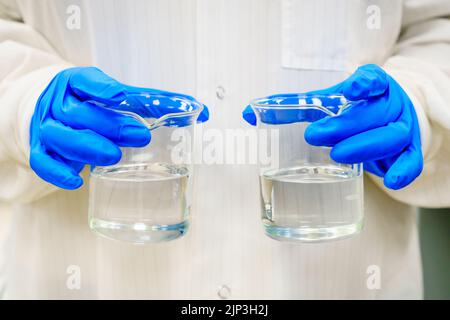 Eine Person in einem Labormantel hält zwei Becher mit klaren Flüssigkeiten Stockfoto