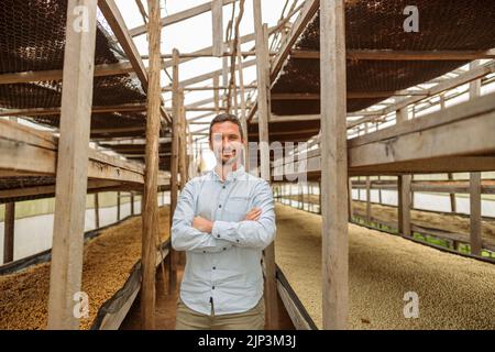 Glücklicher Mann, der zwischen hohen Holzregalen auf der Kaffeefarm steht Stockfoto