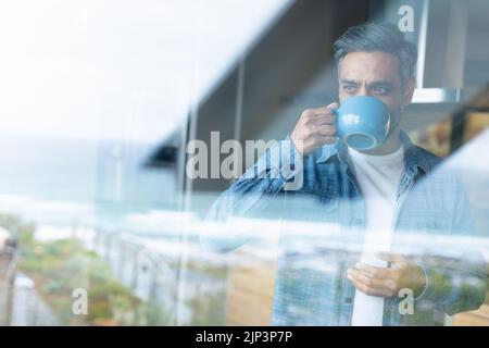 Glücklicher Birazialmann, der Becher hält und durch das Fenster in der Küche schaut Stockfoto