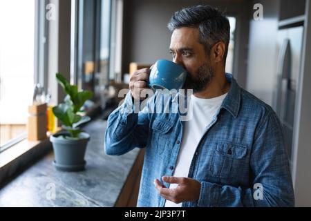 Glücklicher Birazialmann, der Becher hält und durch das Fenster in der Küche schaut Stockfoto