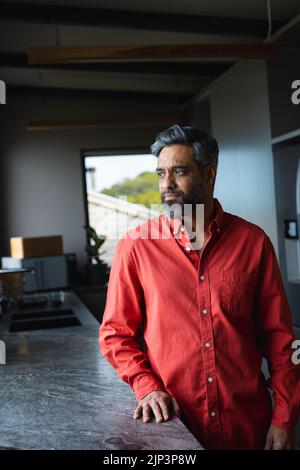 Nachdenklicher Birazialmann, der sich auf der Arbeitsfläche lehnte und durch das Fenster in der Küche schaute Stockfoto