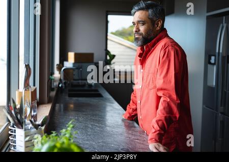 Glücklicher Birazialmann, der sich auf der Arbeitsplatte lehnte und durch das Fenster in der Küche schaute Stockfoto