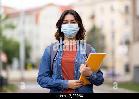 Junge arabische Studentin, die eine medizinische Gesichtsmaske trägt und im Freien posiert Stockfoto