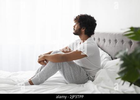 Nachdenklicher indischer Kerl, der zu Hause auf dem Bett sitzt Stockfoto