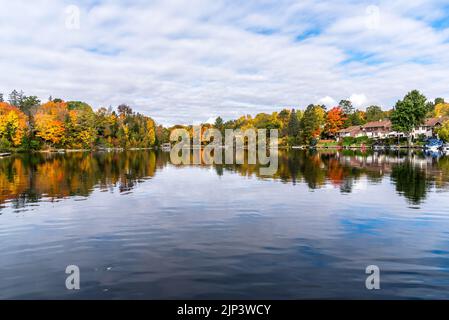 Häuser und Bootshäuser an den bewaldeten Ufern eines Sees an einem bewölkten Herbstmorgen. Atemberaubende Herbstfärbung und Spiegelung in den ruhigen Gewässern. Stockfoto